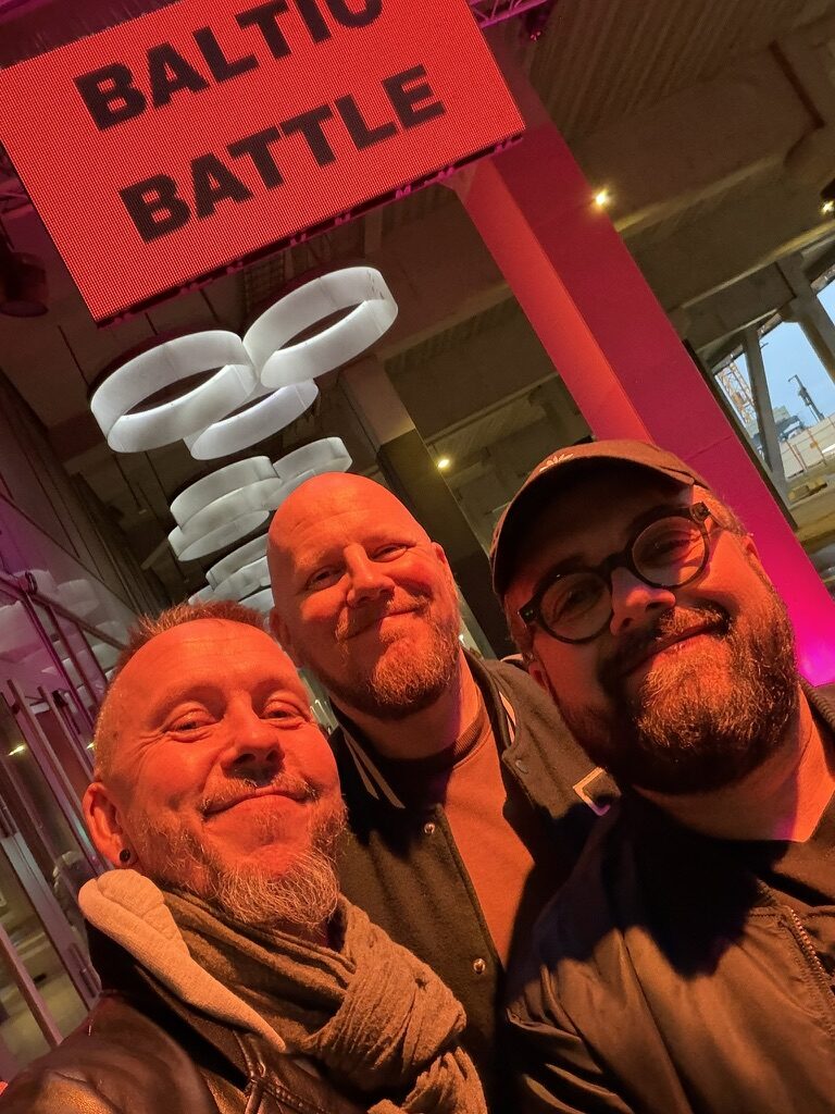 Three smiling men posing for a selfie outside A75 under the glowing red Baltic Battle sign